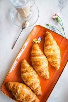 A delightful breakfast with croissants and coffee on a marble table. Croissants are golden brown on an orange plate, coffee with froth.