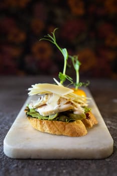 A bruschetta with chicken, pesto, and parmesan on a white marble board. The bruschetta is garnished with microgreens and a pea pod.