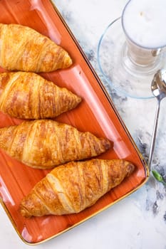 A delightful breakfast with croissants and coffee on a marble table top view. Croissants are golden brown on an orange plate, coffee with froth.