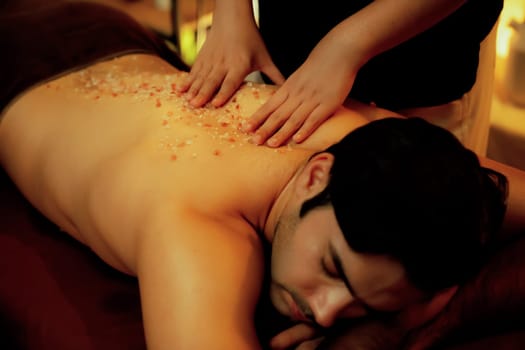 Man customer having exfoliation treatment in luxury spa salon with warmth candle light ambient. Salt scrub beauty treatment in Health spa body scrub. Quiescent