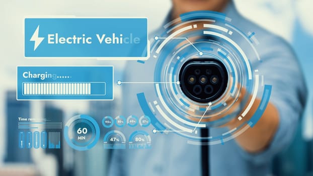 Focus EV charger pointing in front of camera display smart digital battery status hologram with blur cityscape background. EV car charger using alternative clean energy reducing CO2 emission. Peruse