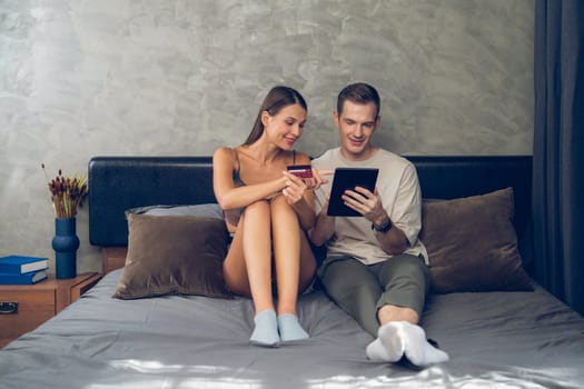 Young couple sit in the home bedroom using online payment app and digital wallet on smartphone to pay with credit card. E commerce shopping and modern purchasing via mobile internet. Unveiling