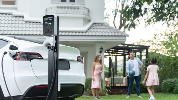 Focus electric vehicle recharge from home charging station on blur background of happy and playful family playing together. EV car using alternative and sustainable energy for better future.Synchronos