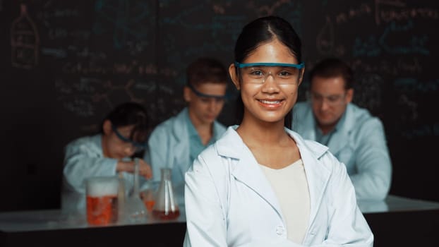 Girl looking at camera with arm folded while people doing experiment at laboratory. Cute student standing blackboard with chemical theory with blurring background at STEM science class. Edification.