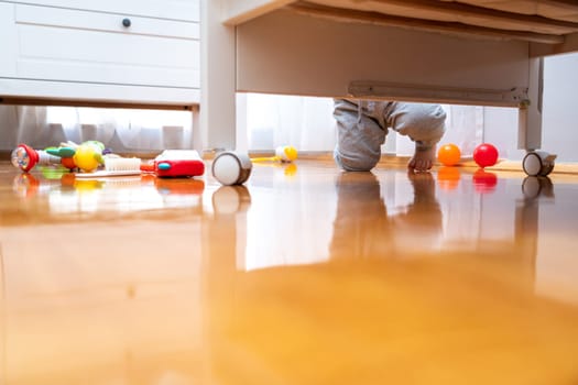 Baby standing near a crib with toys on the floor. View from under the bed. The window brings in