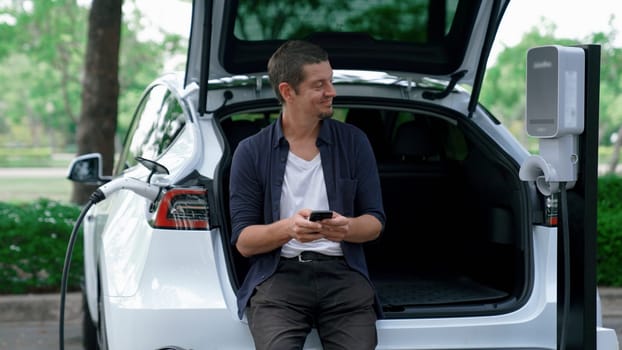 Man using smartphone online banking application to pay for electric car battery charging from EV charging station during vacation holiday road trip at national park or summer forest. Exalt