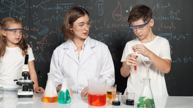 Caucasian boy mixing chemical liquid while teacher giving advice. Professional instructor wearing lab suit looking for diverse student at table with beaker filled with colored solution. Erudition.