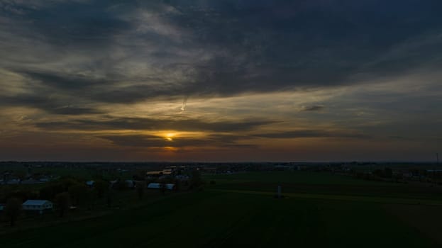 Dramatic Sunset Casting A Warm Glow Over A Rural Landscape With Silhouetted Trees And Buildings Under A Cloud-Streaked Sky.