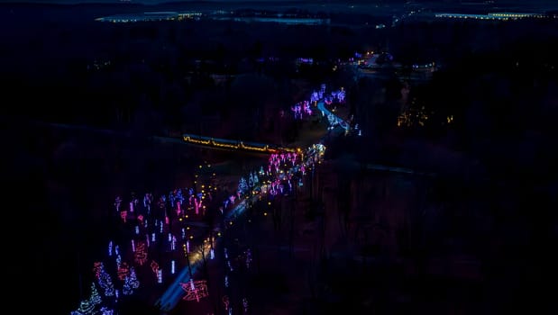 Aerial Night View Capturing A Winding Road Flanked By Colorful Trees With Christmas Lights Leading Towards A Distant Illuminated Area.