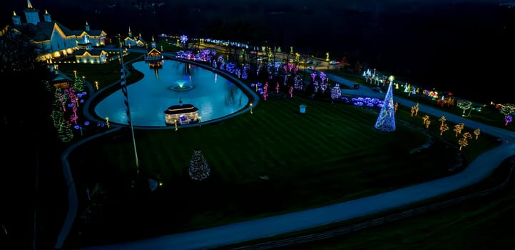 Aerial View Of A Reflective Pond Amidst A Serpentine Pathway Surrounded By Buildings And Trees Adorned With Vibrant Holiday Lights At Night.