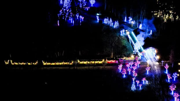 Aerial View At Night Capturing The Glow Of Colorful Lights On Trees Alongside A Smoke-Emitting Train Moving Through A Dark Forest.