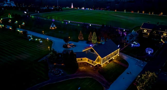 Aerial View Of An Evening Scene With Illuminated Buildings And A Large Field Surrounded By Christmas Trees And Light Decorations.