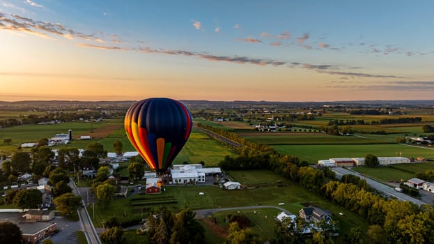 Colorful Hot Air Balloon Floating Above A Scenic Rural Landscape With Farms, Fields, And A Small Town During Sunset.