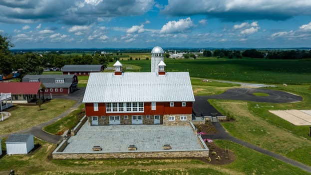 Overhead View Of A Large Red And Gray Barn With A Shiny Metal Roof, Accompanied By Smaller Buildings, Nestled In A Rural Setting With Lush Fields Extending To The Horizon Under A Partially Cloudy Sky.