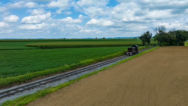 Single Locomotive Traversing Along Railway Tracks Bordering A Plowed Field And Green Crops Under A Vast Sky With Puffy Clouds.