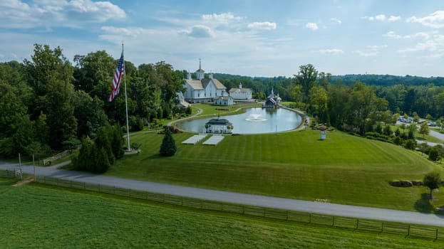 Aerial View Of A Grand White Building With A Grey Roof Surrounded By Lush Green Lawns, A Large Reflective Pond With Fountains, And A Majestic Flagpole Bearing The American Flag.