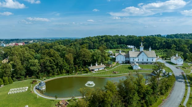 Expansive Aerial View Over A Religious Complex With Multiple White Buildings And Shining Golden Domes, A Pond With A Fountain, Set In A Verdant Landscape With A Large American Flag, With Fluffy Clouds