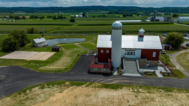 Overlooking A Farmstead Featuring A Majestic Red Barn With Silo, An Adjacent Pond With Geese, Winding Asphalt Paths, And Lush Green Crop Fields Under A Blue Sky With White Clouds.