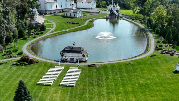 Aerial View Showcasing A Cluster Of Traditional White Orthodox Churches With Cross-Topped Domes, Arranged Around A Curved Pond With A Fountain, Amidst Green Trees And Neatly Arranged White Benches