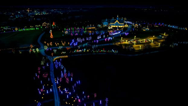 High-Angle Night View Of A Lively Holiday Lighting Display With A Road Leading To A Central Pond And Clustered Lit Structures.