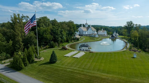 Aerial View Of A Grand White Building With A Grey Roof Surrounded By Lush Green Lawns, A Large Reflective Pond With Fountains, And A Majestic Flagpole Bearing The American Flag.