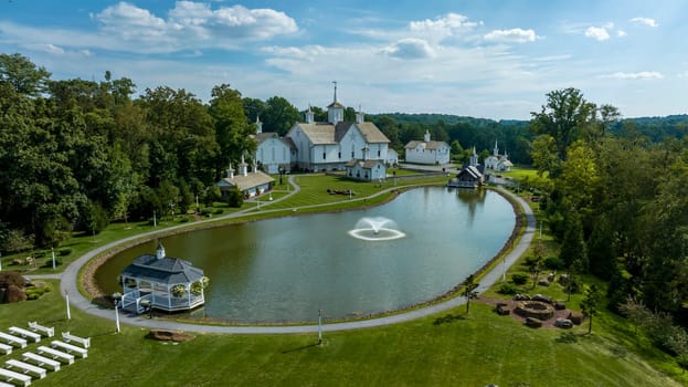 Aerial View Showcasing A Cluster Of Traditional White Orthodox Churches With Cross-Topped Domes, Arranged Around A Curved Pond With A Fountain, Amidst Green Trees And Neatly Arranged White Benches