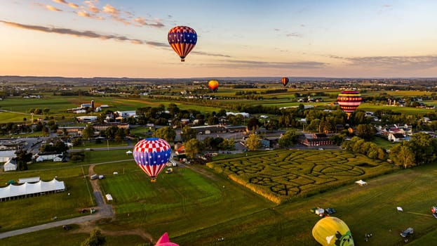 An Aerial View of Multiple Hot Air Balloons Soar Over A Picturesque Landscape Featuring A Corn Maze, Town, And Fields During A Golden Hour Sunset.