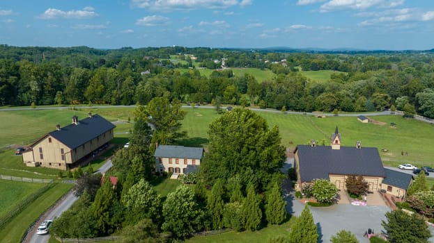 Scenic Overlook Of A Rural Community With Multiple Large Buildings, Including A Church, Amidst Well-Maintained Grass Fields, Enclosed By Wooden Fences, With Dense Forests In The Background