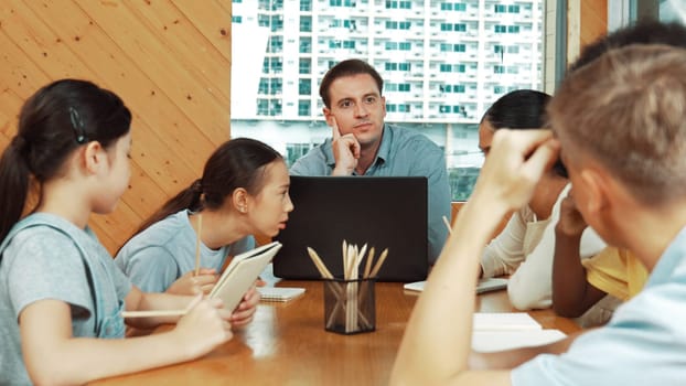 Caucasian teacher and smart boy discuss about project at classroom. Professional instructor talking about classwork while giving a paper to highschool child with diverse student listen. Edification.