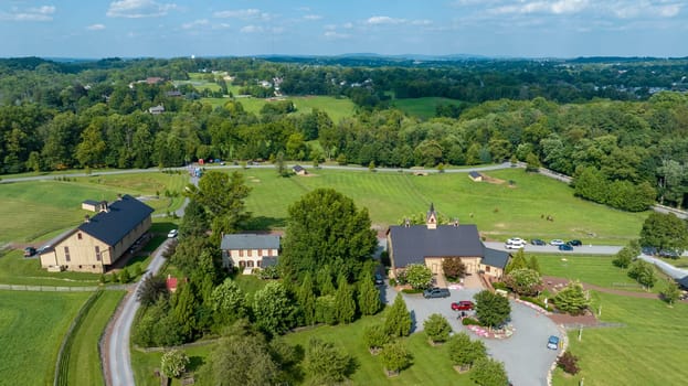 Aerial View Over A Pastoral Setting With A Curving Driveway Leading To Buildings Amidst Large Green Fields, Enclosed By Wooden Fences, With Distant Horizon Of Trees And Hills Under Cloud-Speckled Sky.