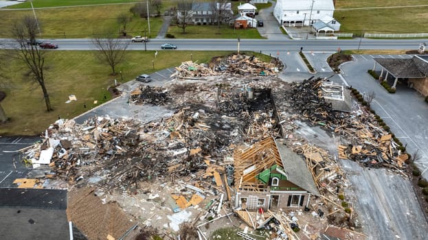 An Aerial View Of A Devastated Building Amidst Intact Structures With Cars On The Road And Green Fields In The Background Under A Cloudy Sky.