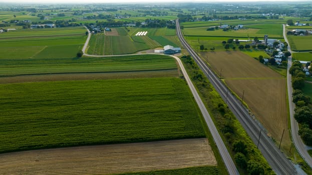 High Angle View Showcasing A Stretch Of Road And Railroad Tracks Cutting Through A Lush Tapestry Of Agricultural Fields With Farm Buildings In The Distance.