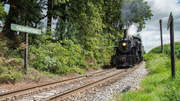 Black Steam Locomotive Number 89 Blowing Steam On Rural Tracks Next To 'Carpenters' Sign Surrounded By Verdant Foliage.