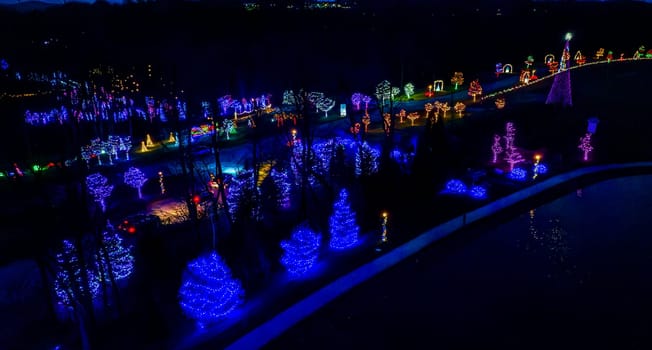 Night View Of A Vibrant Display Of Blue And Purple Christmas Lights On Trees Lining A Road With Cars And A Distant Area Aglow.