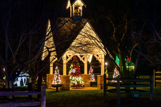 Illuminated Night View Of A Small Chapel Surrounded By Multiple Christmas Trees Decorated With Lights, Visible Through Bare Tree Branches And A Rustic Wooden Fence.