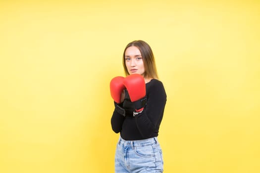 Portrait of young woman in a casual clothes and hands in boxing gloves against yellow background