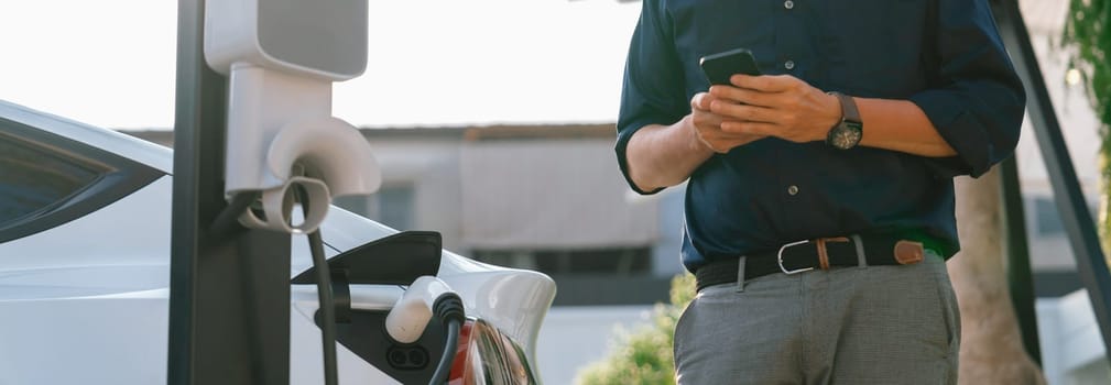 Young man use smartphone to pay for electricity at public EV car charging station at city commercial mall parking lot. Modern environmental and sustainable urban lifestyle with EV vehicle. Expedient