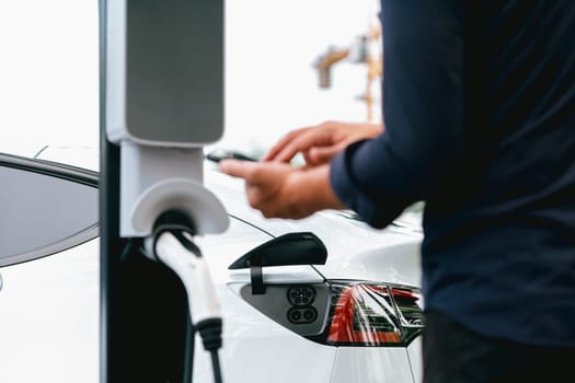 Man using smartphone online banking application to pay for electric car battery charging from EV charging station during vacation holiday road trip at national park or summer forest. Exalt