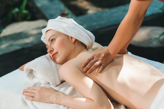 Beautiful young woman received a back massage on a spa bed from professional masseuse. Attractive female relaxes deeply by skilled hands of the massage therapist. Surrounded with nature. Tranquility.