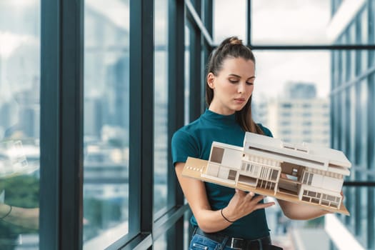 Beautiful young architect engineer holds architectural model while inspect house model. Professional interior designer checking house construction while standing near window with city view. Tracery.