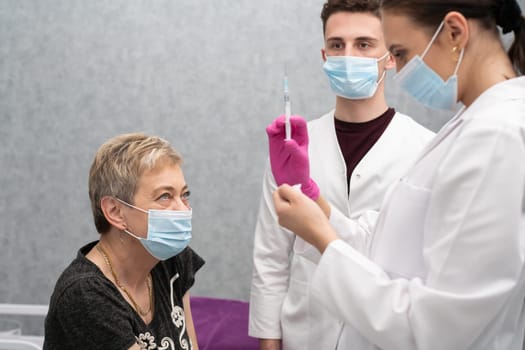 Before the next vaccine, the doctor checks the amount and the contents in the syringe. A woman in her 50s is sitting waiting for an injection. The medical intern observes and learns all activities during the time of vaccination of persons.