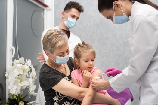 Grandma took her granddaughter on her lap so that the child would not be afraid of the injection made by the young nurse. A young nurse is injecting a new vaccine. Safe vaccination of children.