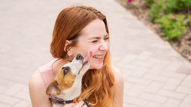 Dog jack russell terrier licks the owner in the face outdoors. Girl with braces on her teeth