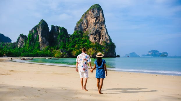 a diverse couple of men and women on the beach, an Asian woman and a caucasian man walking on the beach of Railay Beach in Thailand with limestone cliffs in the background at sunrise