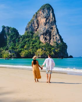 a diverse couple of men and women on the beach, an Asian woman and a caucasian man walking on the beach of Railay Beach in Thailand with limestone cliffs in the background on a sunny day
