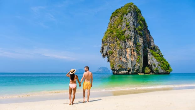a diverse couple of men and women on the beach, an Asian woman in bikini and a caucasian man in swim short walking on the beach of Railay Beach in Thailand with limestone cliffs in the background