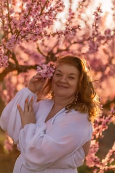 Woman blooming peach orchard. Against the backdrop of a picturesque peach orchard, a woman in a long white dress enjoys a peaceful walk in the park, surrounded by the beauty of nature