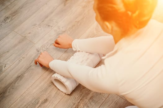 Adult athletic woman, in white bodysuit, performing fascia exercises on the floor - caucasian woman using a massage foam roller - a tool to relieve tension in the back and relieve muscle pain - the concept of physiotherapy and stretching training.