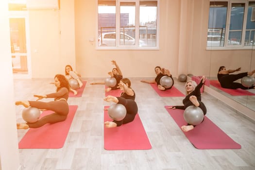 A group of six athletic women doing pilates or yoga on pink mats in front of a window in a beige loft studio interior. Teamwork, good mood and healthy lifestyle concept