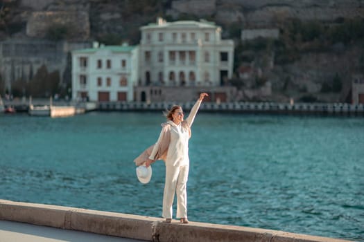 Happy blonde woman in a white suit and hat posing at the camera against the backdrop of the sea.